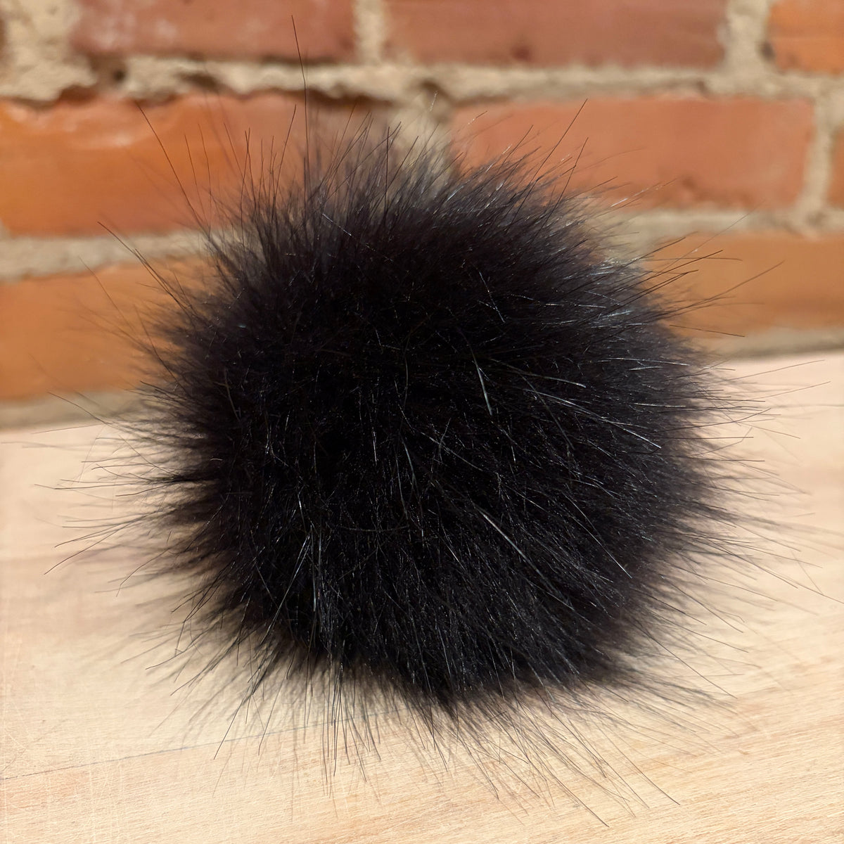 A fluffy black faux fur pom-pom ball resting on a light wood surface with a brick wall background.