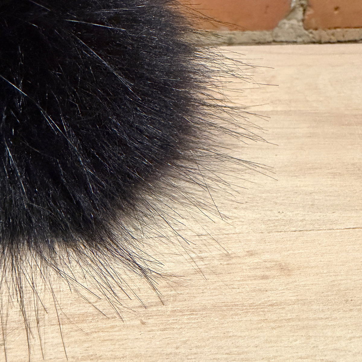 A close-up of a fluffy black faux fur pom pom resting on a light-colored wooden surface with a brick wall in the background.