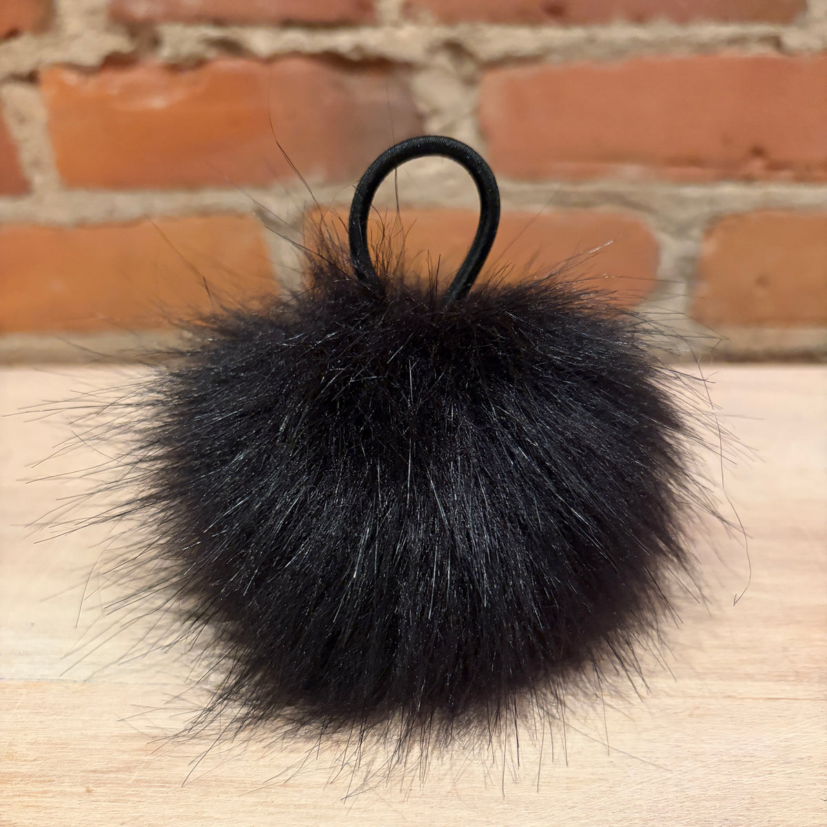 A black faux fur pom-pom keychain or bag charm with a black elastic loop, resting on a light wood surface with a blurred brick wall background.