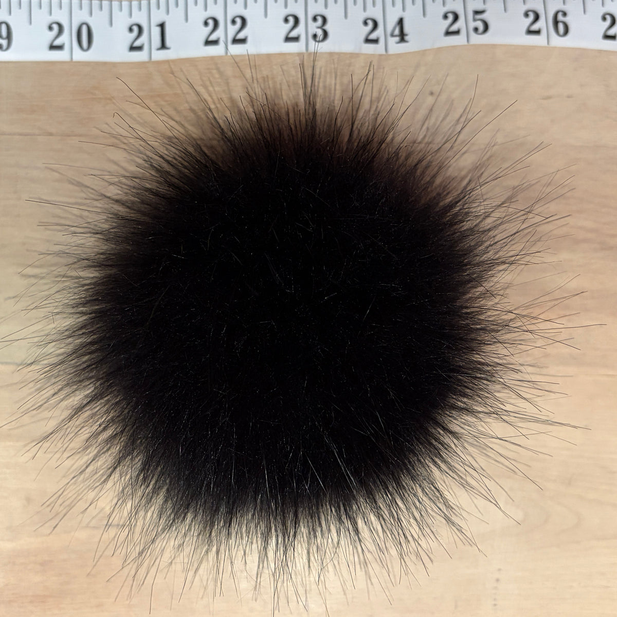 A round, fluffy black faux fur pom-pom or decorative ball shown from above on a light wood surface, with a measuring tape visible along the top edge for scale.