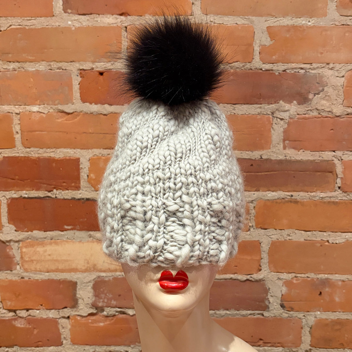A chunky knit beanie hat in a light gray and white variegated yarn, topped with a large black faux fur pom-pom, displayed on a mannequin head against a red brick wall background.