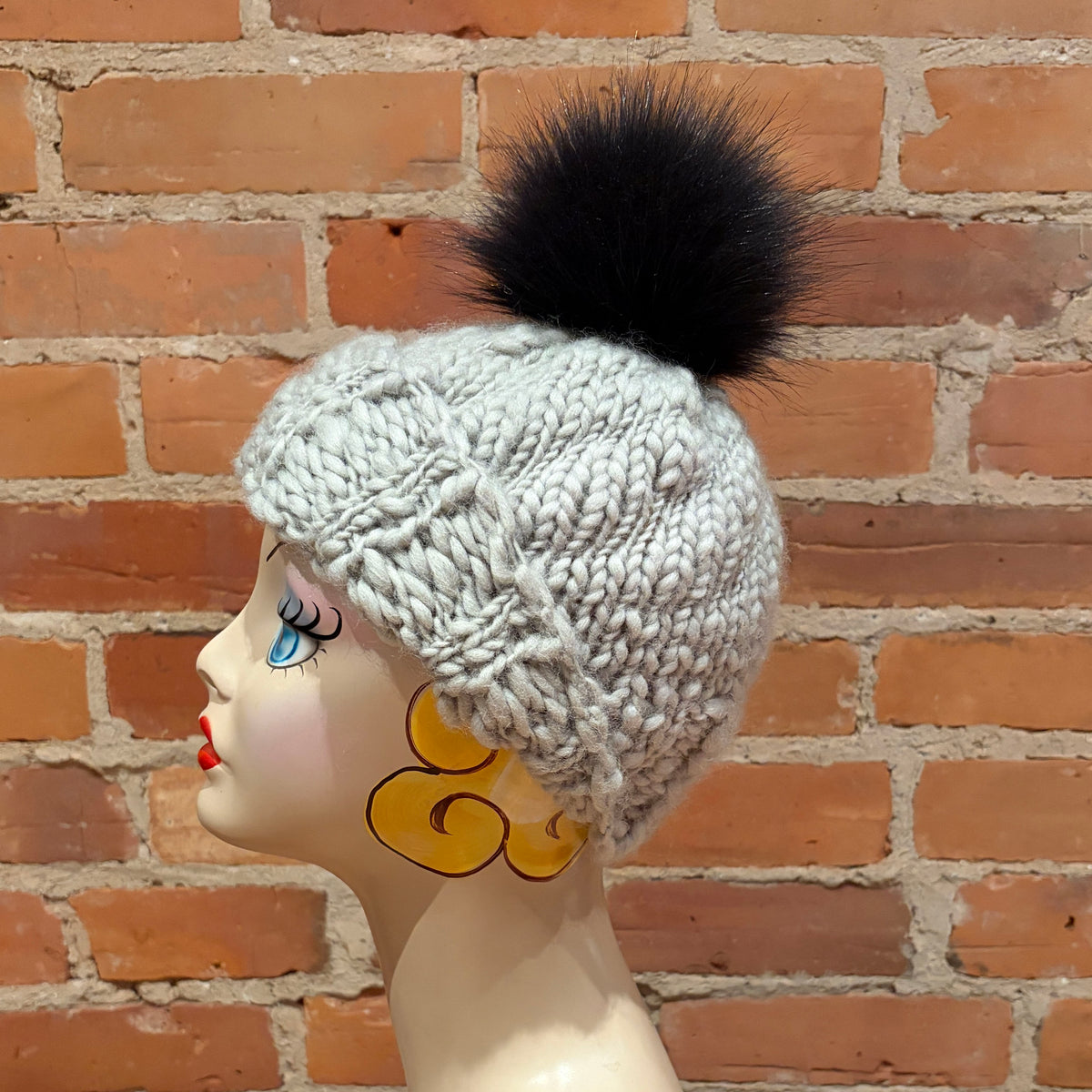 A light gray chunky knit beanie with a large black faux-fur pom-pom, displayed on a mannequin head in profile against a red-brown brick wall.