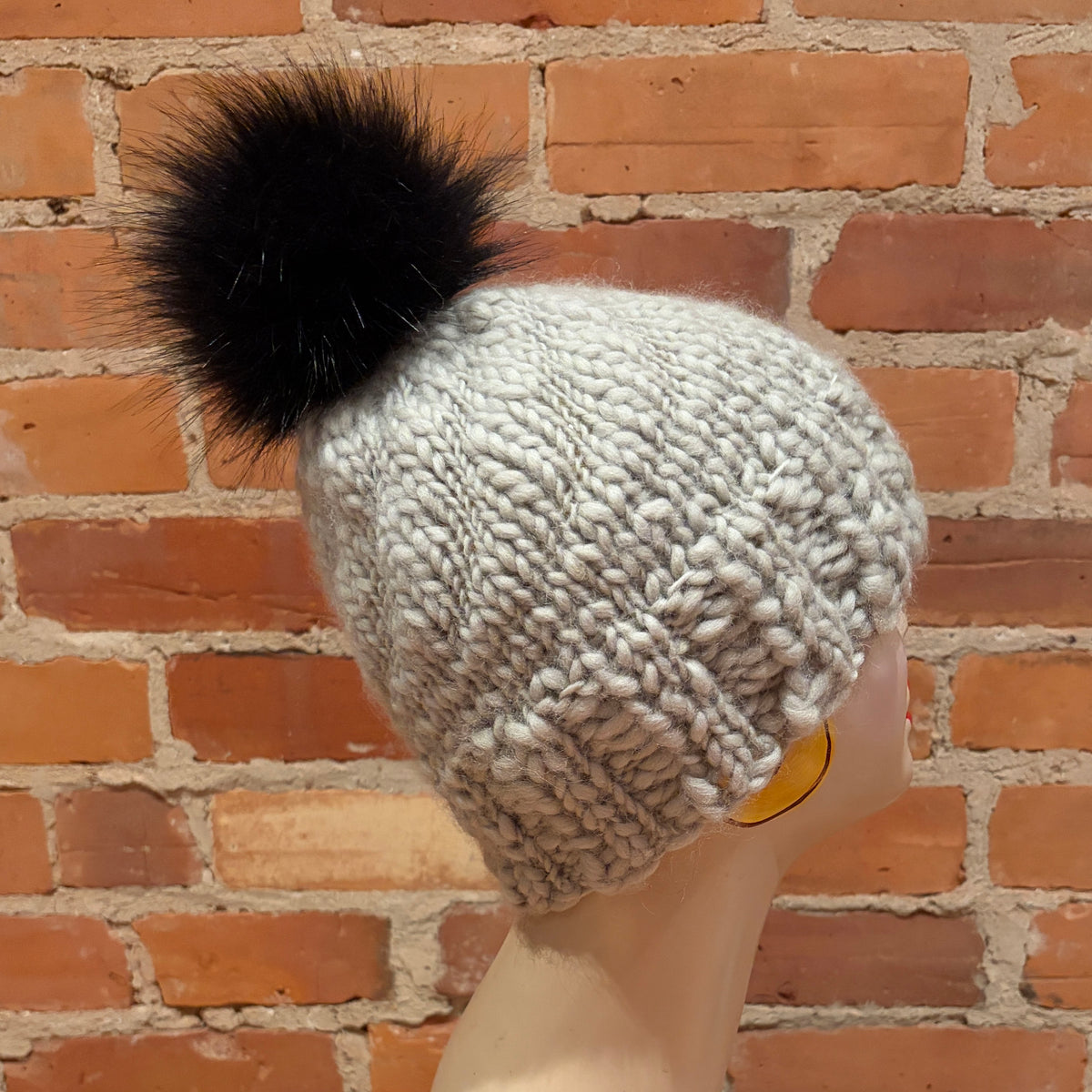 A chunky knit beanie hat in a light oatmeal gray color with a large black faux fur pom-pom, shown on a mannequin head against a red brick wall background.