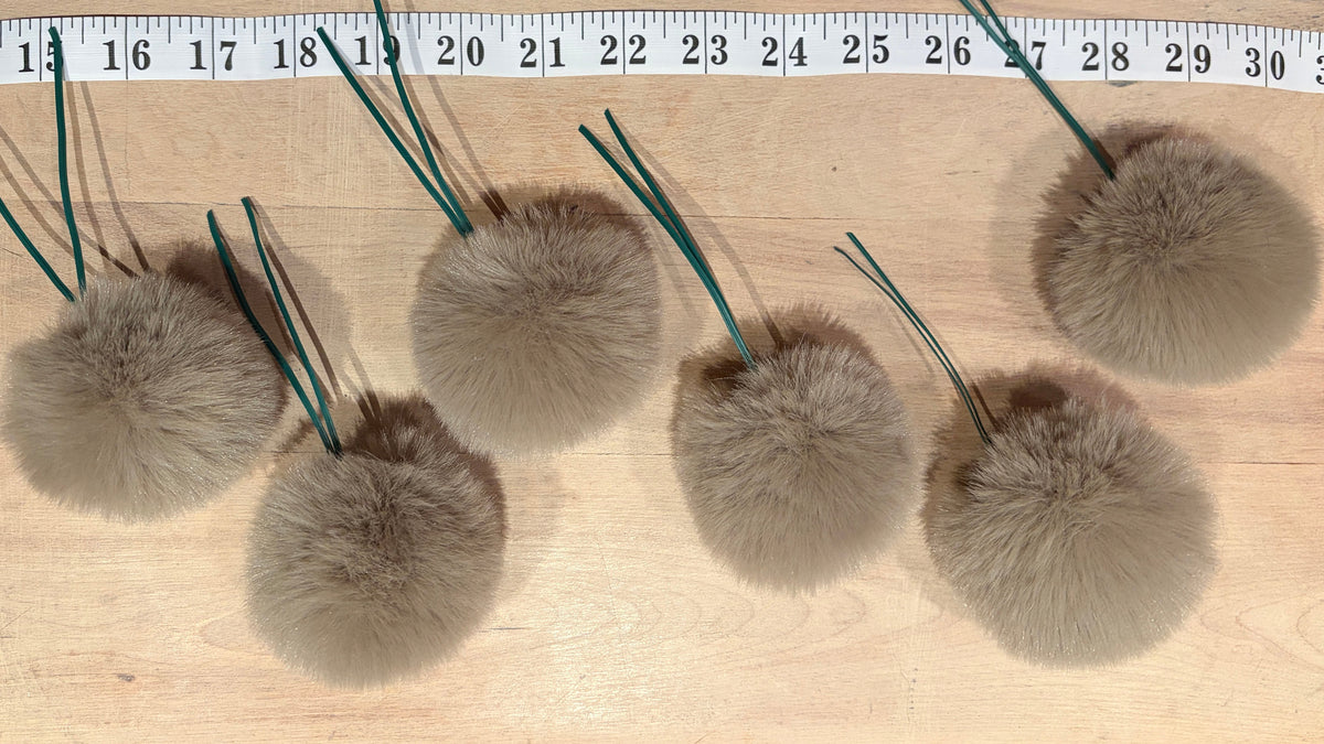 Five light brown faux fur pom-poms with green wire loops, arranged on a light wood surface next to a measuring tape.