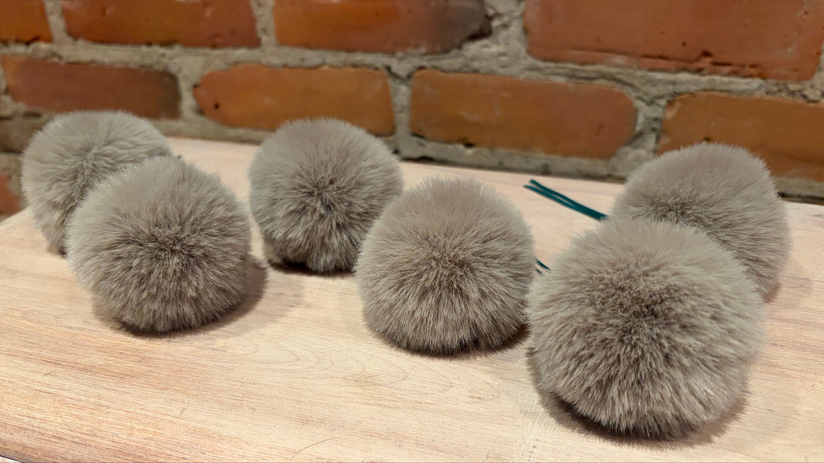 Six fluffy gray faux-fur pom-poms arranged on a light wood surface in front of a brick wall background.
