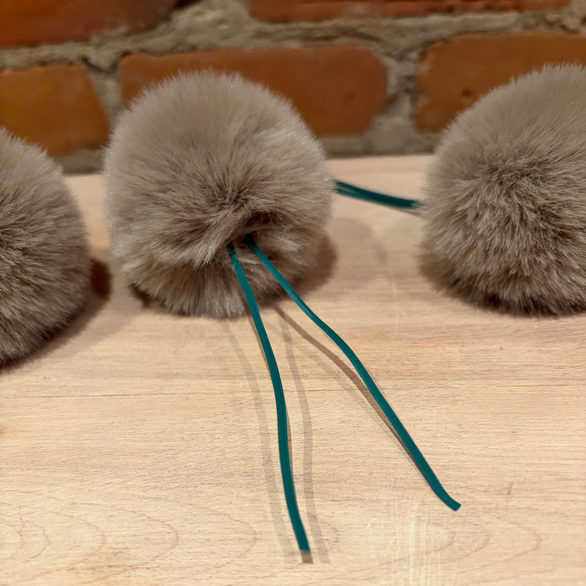 Three fluffy pom-poms in a light taupe-gray color with teal drawstring cords, arranged on a light wood surface with a brick wall in the background.