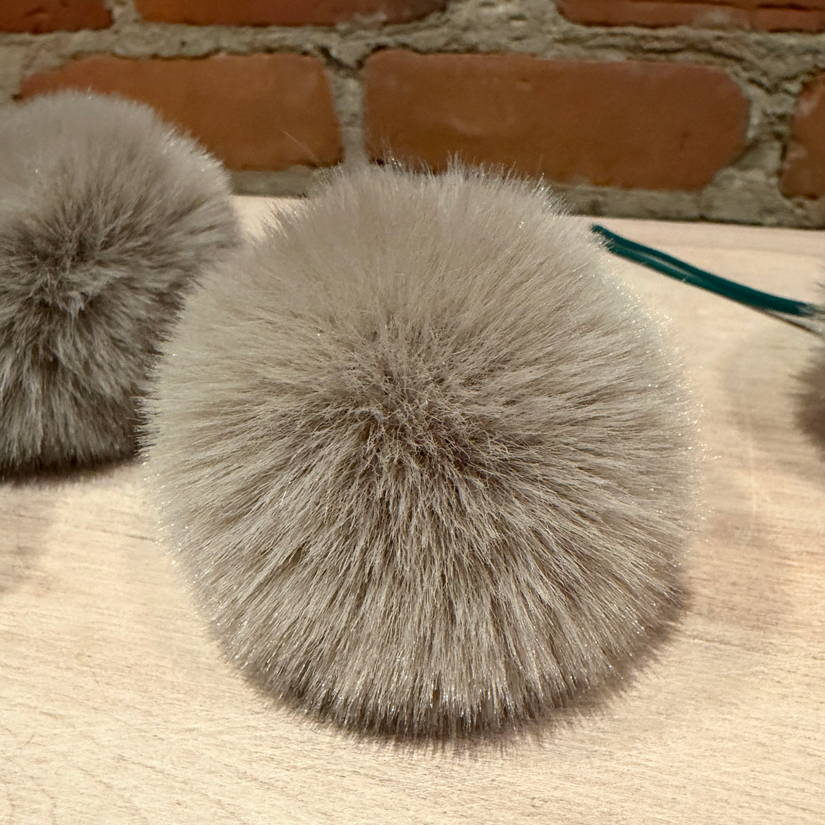 A close-up of a fluffy gray faux fur pom-pom on a light wood surface with a brick wall background.