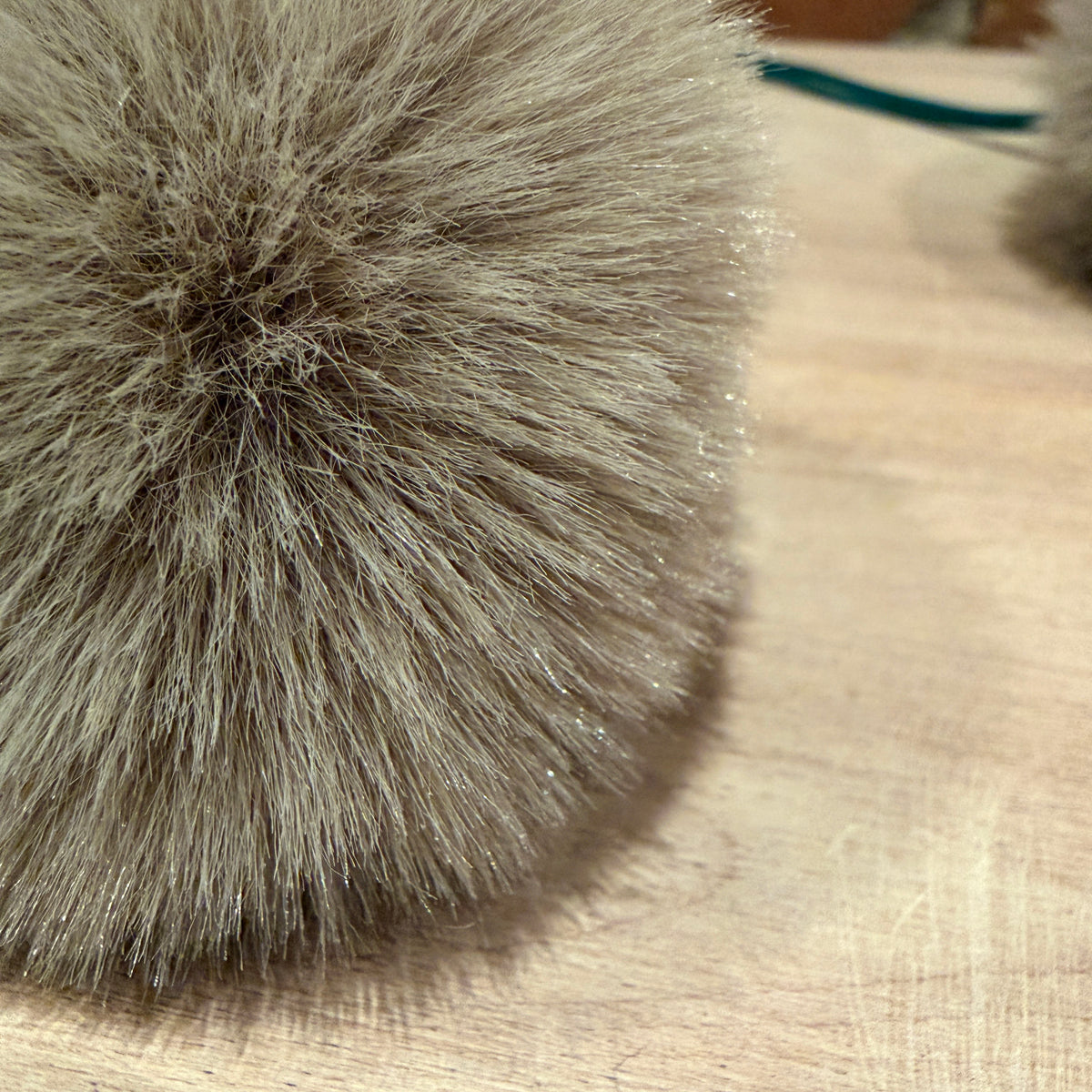 A close-up of a fluffy faux fur pom-pom in a light taupe-gray color resting on a light wood surface.
