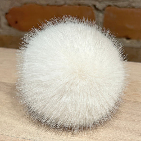 A fluffy white faux fur pom-pom accessory resting on a light wood surface with a brick wall background.