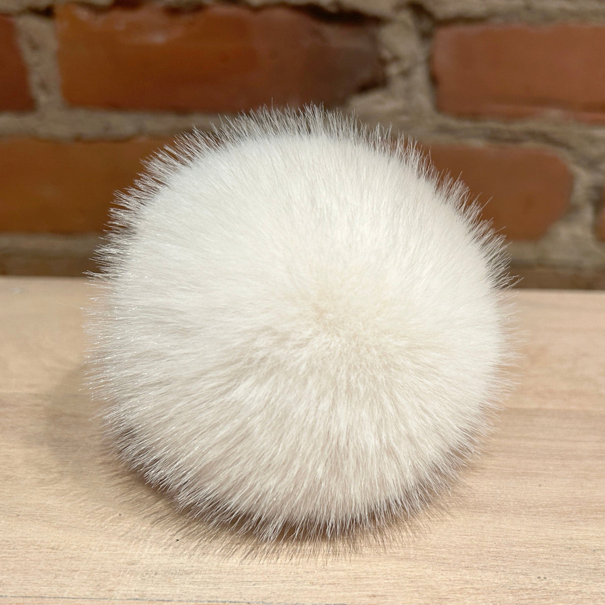 A fluffy white faux fur pom-pom ball resting on a light wood surface with a blurred brick wall background.