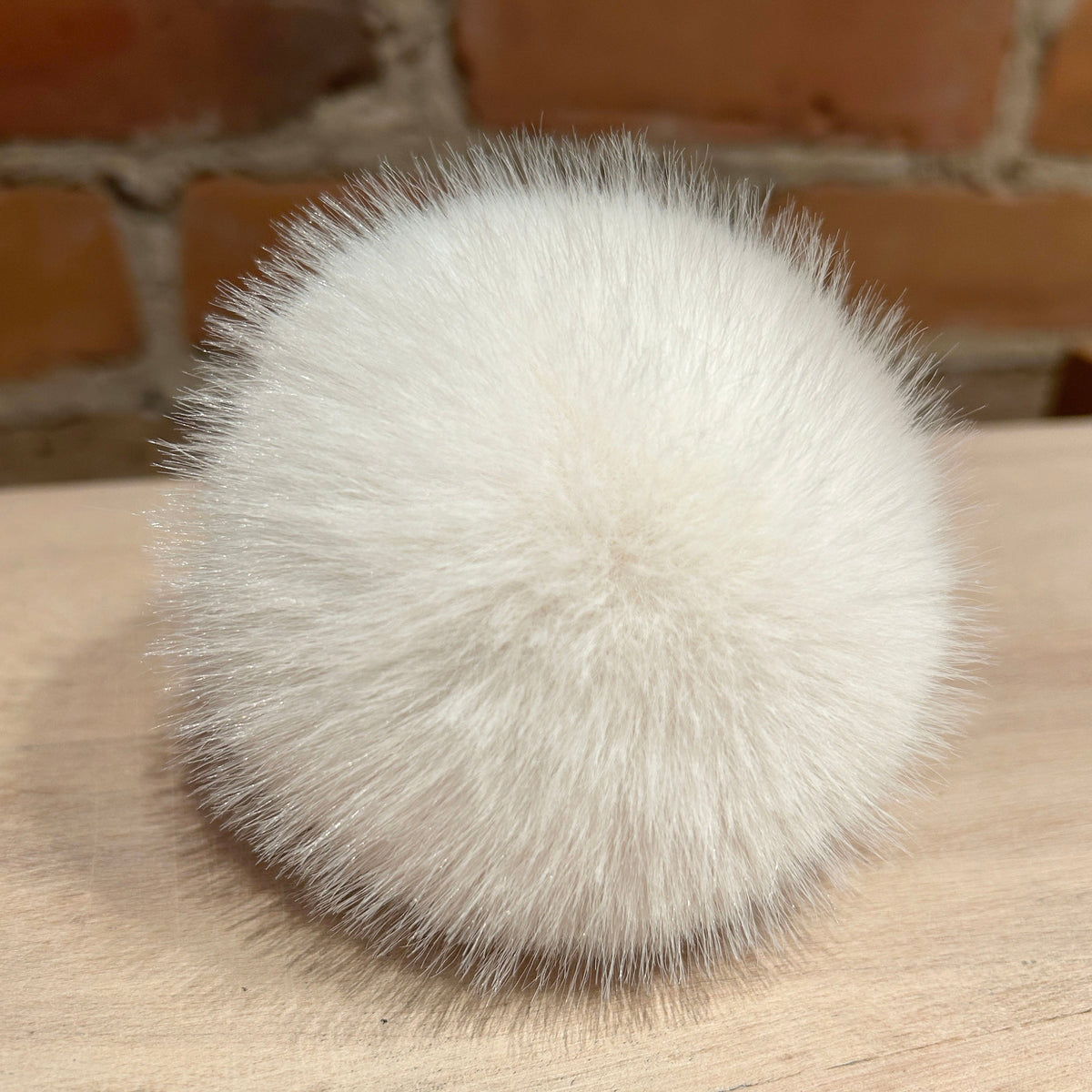 A fluffy pom-pom ball in off-white faux fur or sheepskin, resting on a light wood surface with a blurred brick wall background.