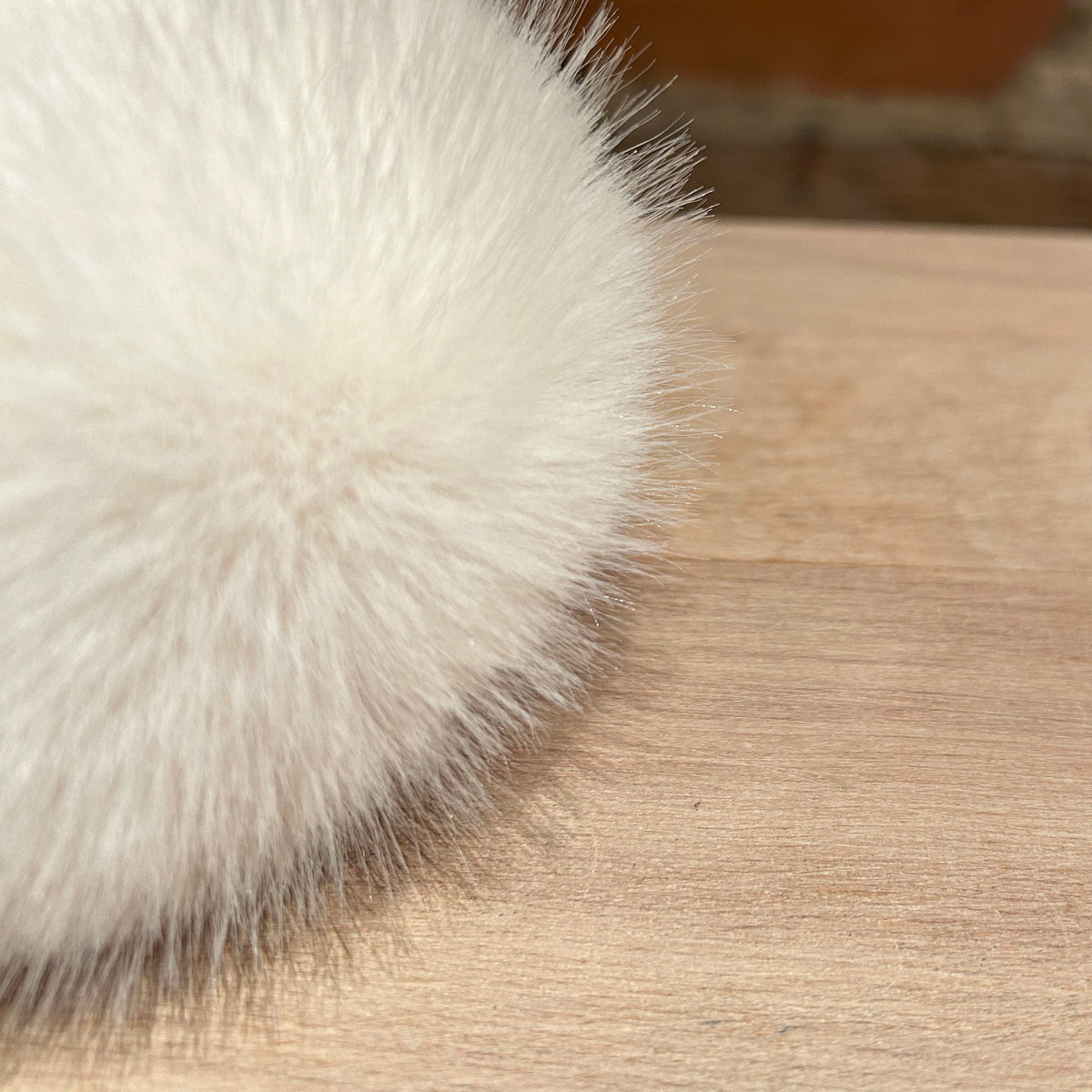 A close-up of a fluffy white faux fur pom-pom resting on a light wood surface with a blurred brick wall background.