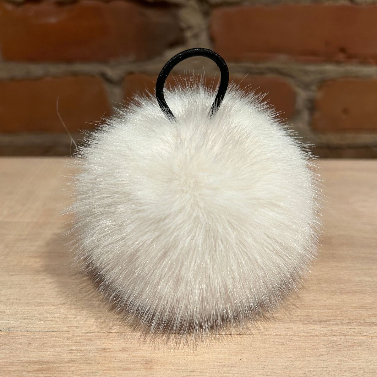 A fluffy white faux fur pom-pom keychain or bag charm with a black elastic loop, resting on a light wood surface with a brick wall background.