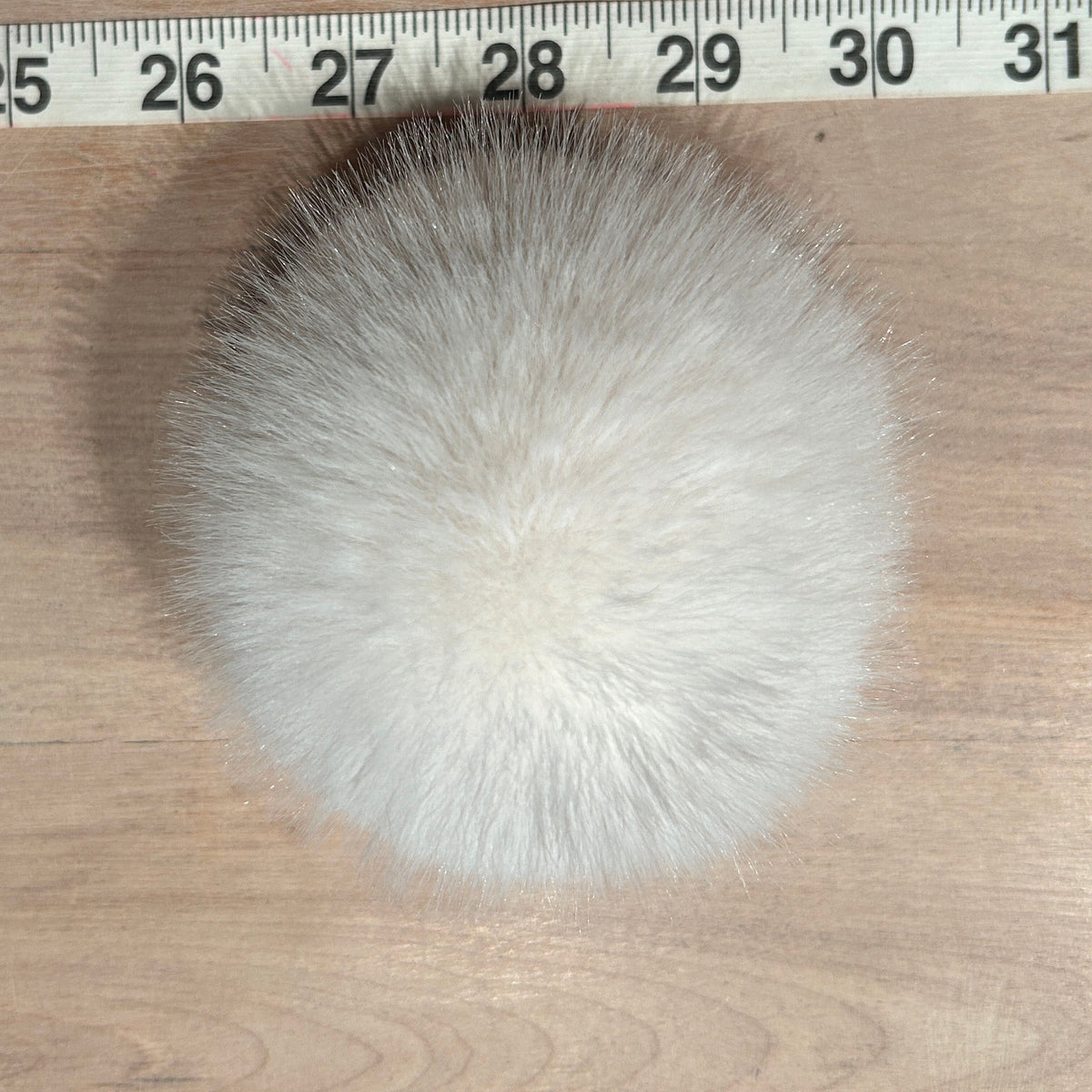 A round fluffy faux fur pom-pom in white and light gray, shown on a wooden surface with a measuring tape ruler at the top for scale.