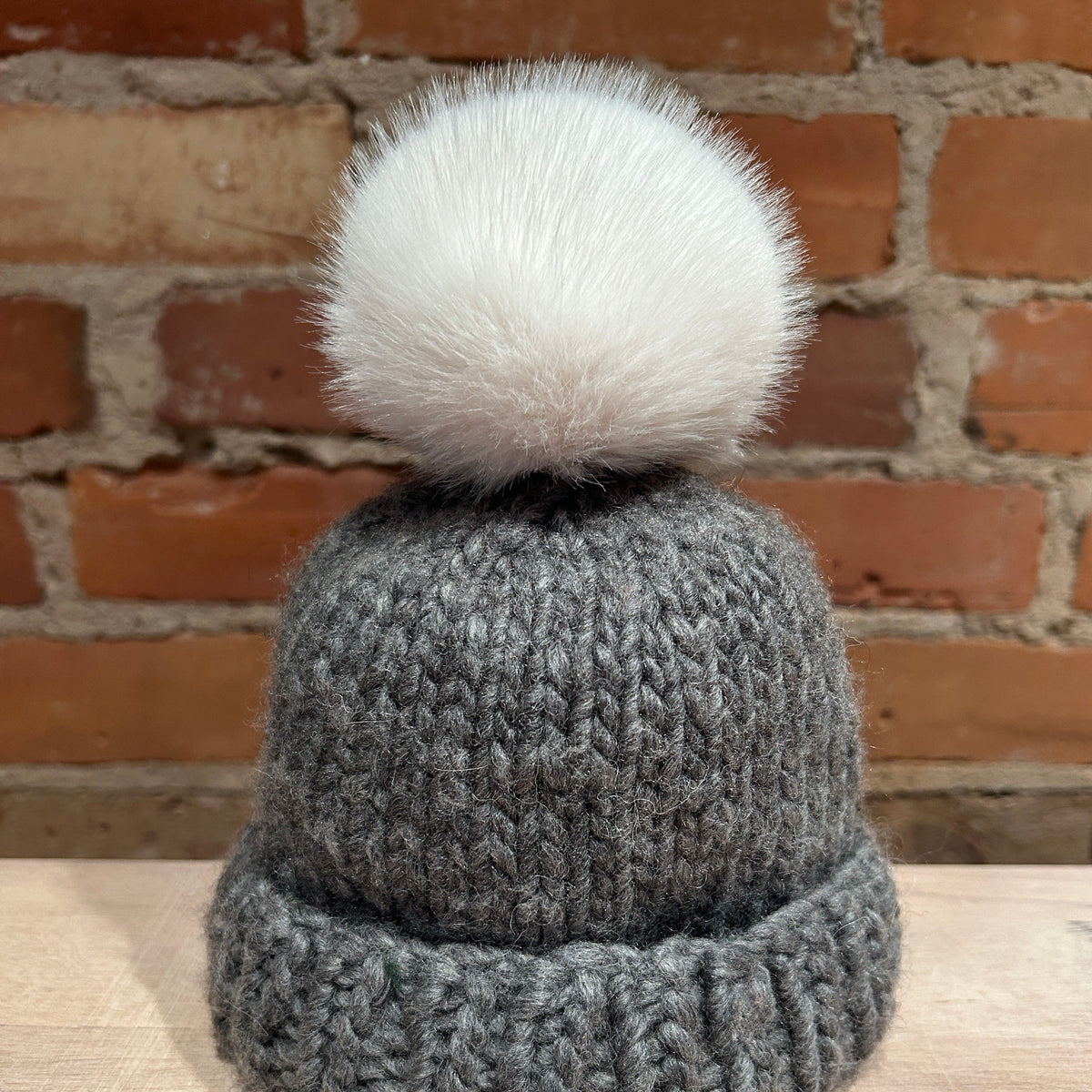 A gray knit beanie hat with a large fluffy white pom-pom, displayed on a light wooden surface against a brick wall background.