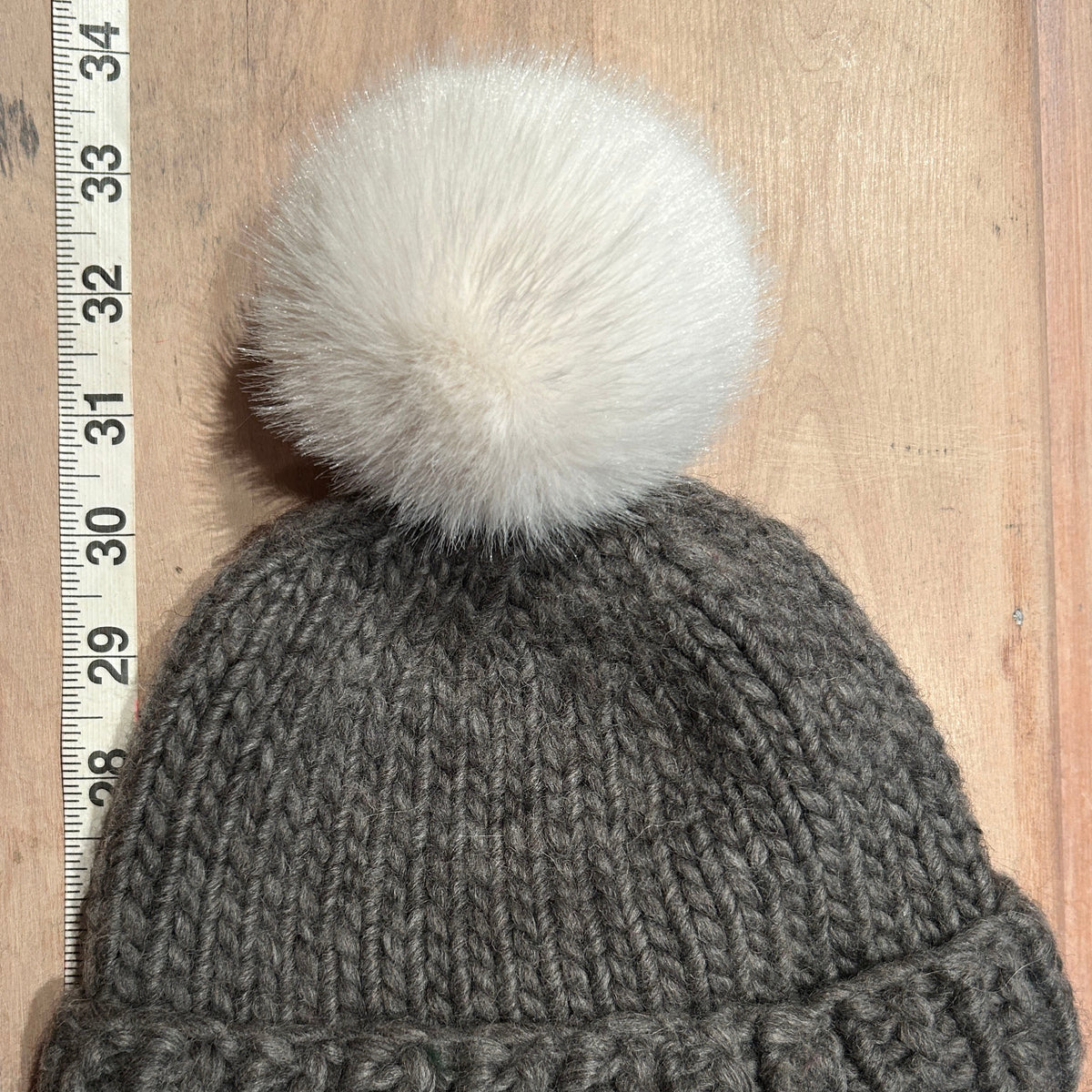 A dark gray knit beanie with a large fluffy white pom-pom, laid flat on a light wood surface next to a white measuring tape ruler.