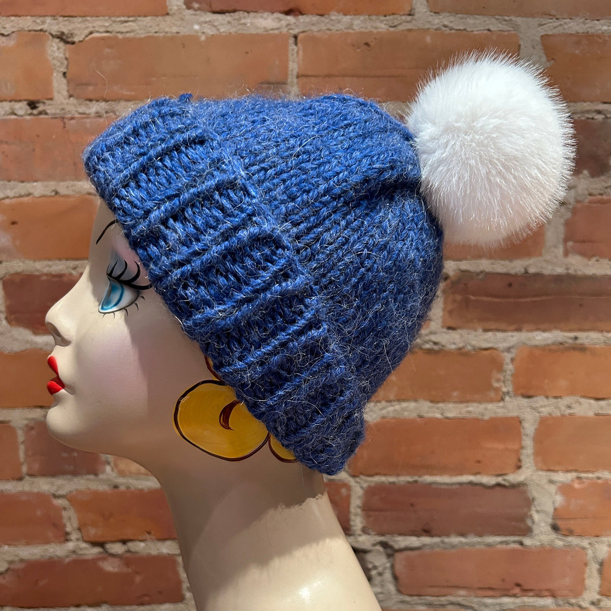 A blue ribbed knit beanie with a fluffy white pom-pom, displayed on a mannequin head against a red brick wall background.