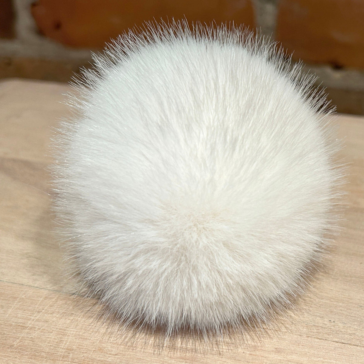 A round, fluffy white faux-fur pom-pom accessory resting on a light wood surface with a blurred brick wall background.