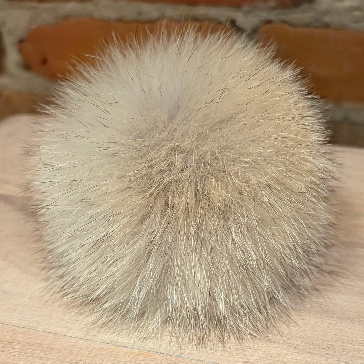 A fluffy coyote-fur pom-pom ball in a light beige and gray color, resting on a light wood surface with a blurred brick wall background.