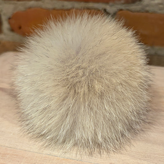 A fluffy coyote-fur pom-pom ball in a light beige and gray color, resting on a light wood surface with a blurred brick wall background.
