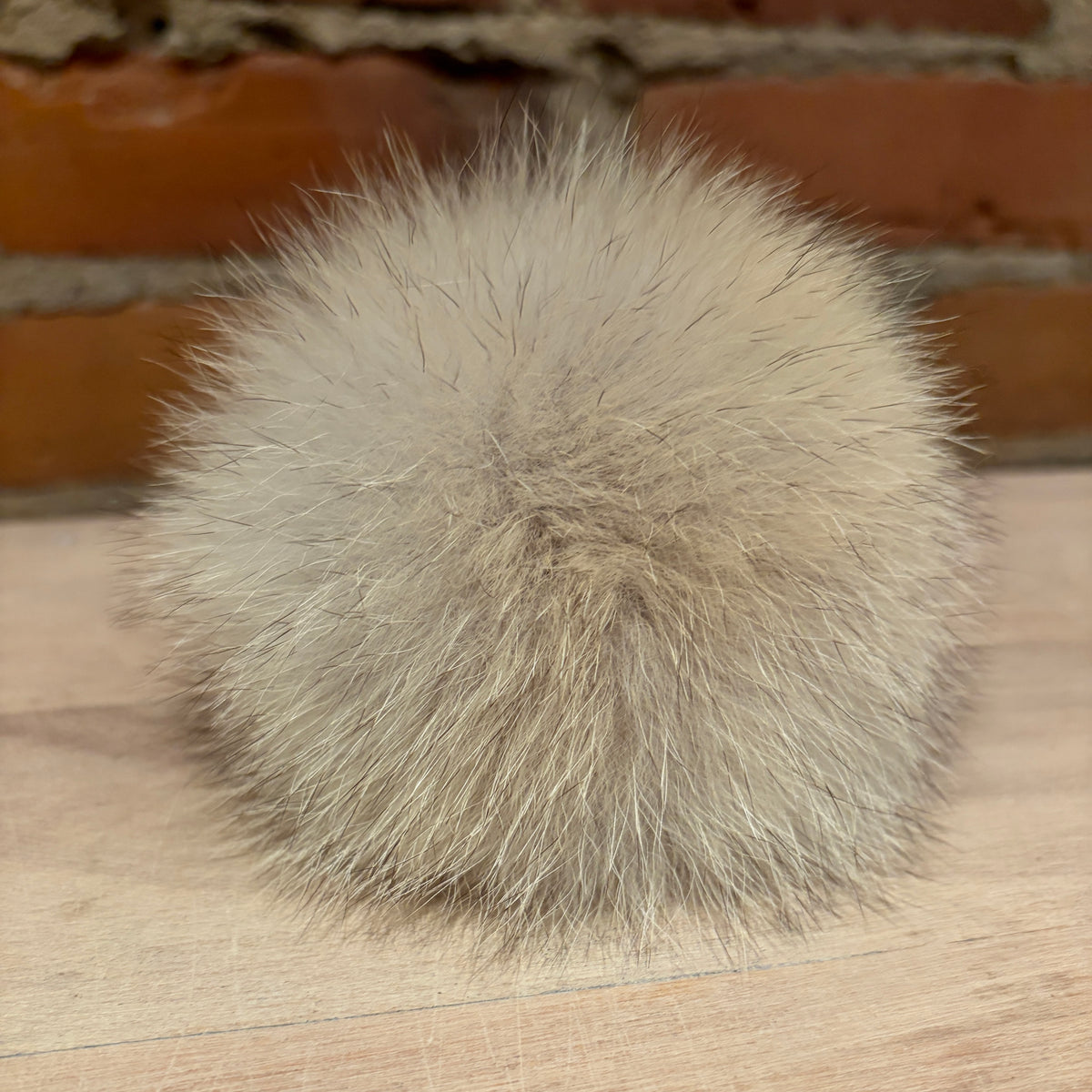 A fluffy pom-pom ball made of light beige and gray coyote fur, resting on a light wood surface with a blurred brick wall background.