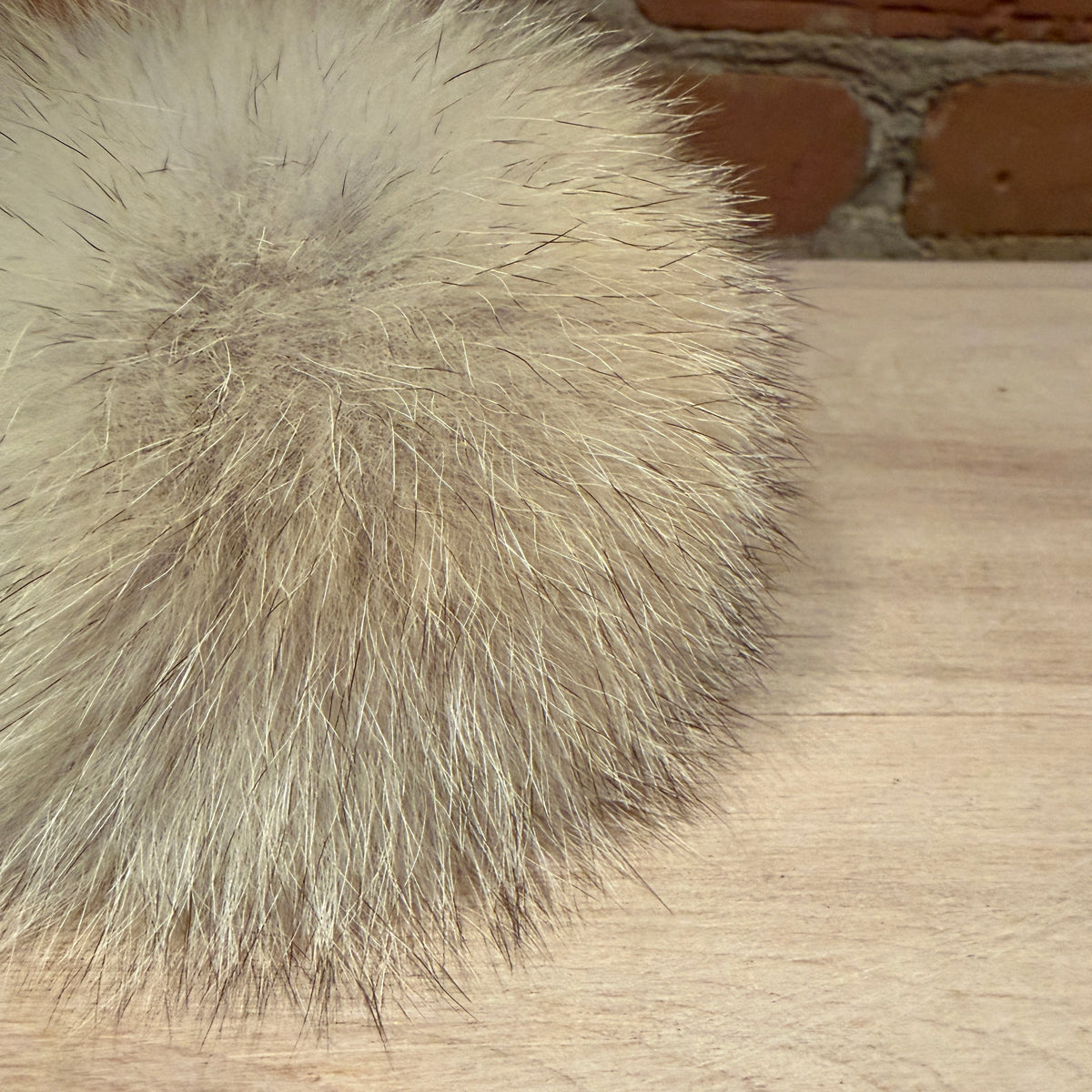 A close-up of a fluffy coyote fur pom-pom in light beige and gray tones, resting on a light wood surface with a brick wall in the background.