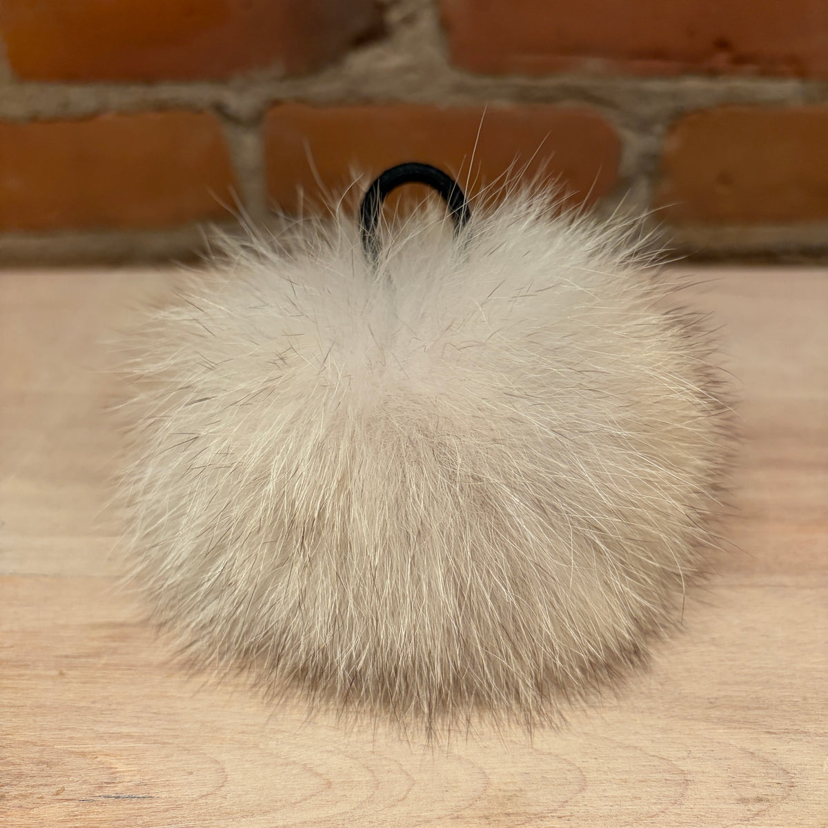 A fluffy pom-pom in a light beige coyote fur material with a black metal ring, resting on a light wood surface with a brick wall background.