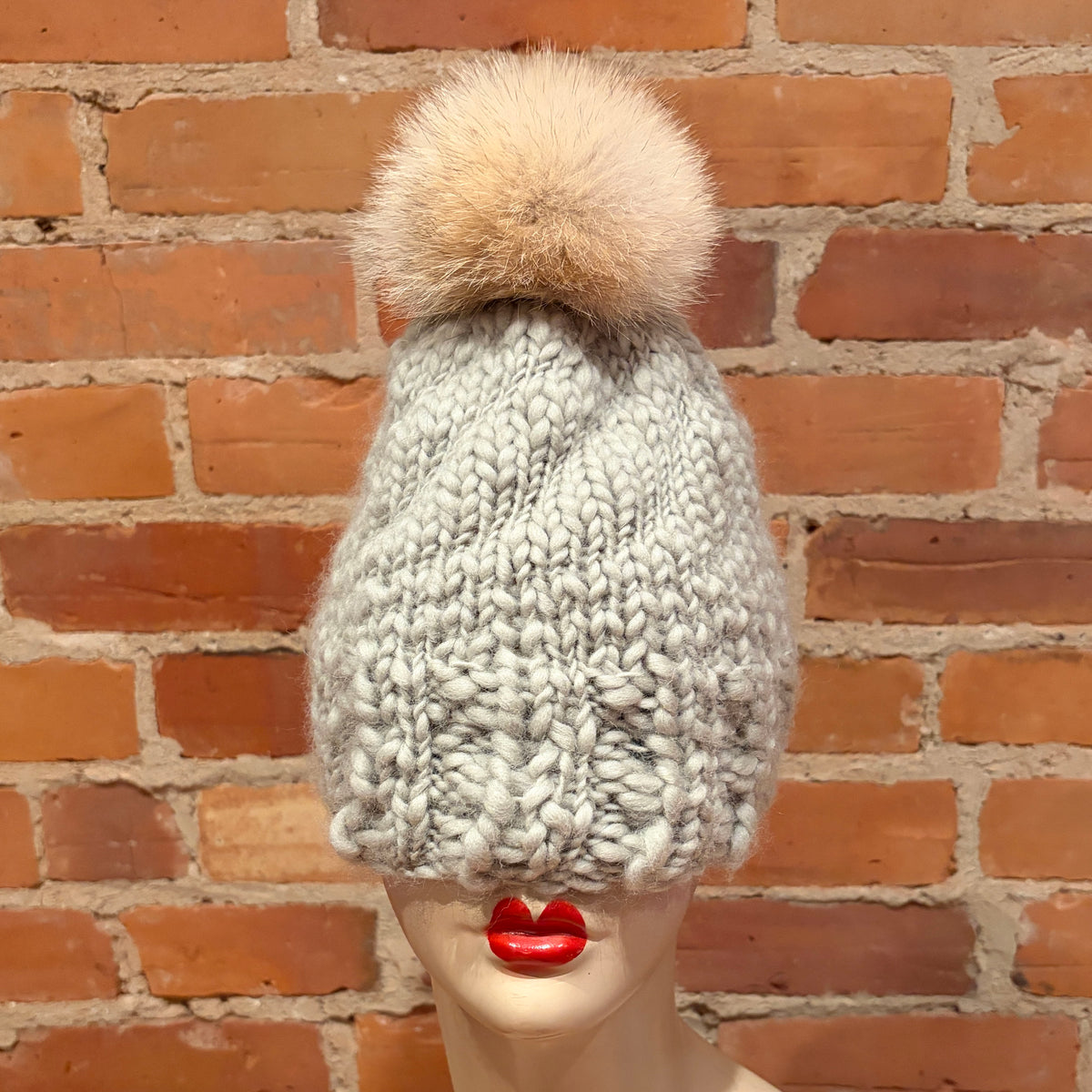 A chunky knit winter beanie in light gray with a large beige coyote fur pom-pom, displayed on a mannequin head against a red brick wall background.