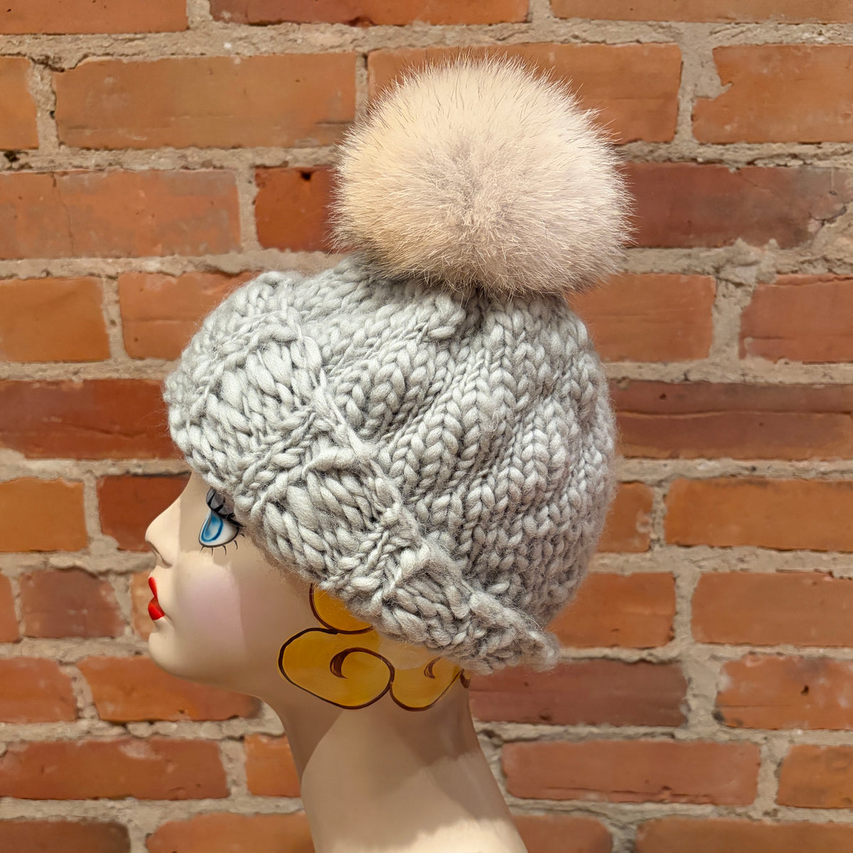 A light gray chunky cable-knit beanie with a fluffy cream pom-pom, displayed on a mannequin head against a red brick wall background.