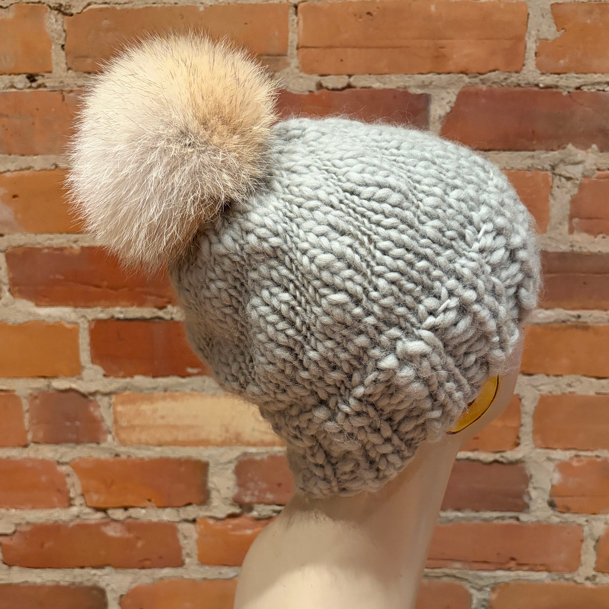 A chunky knit beanie hat in a light gray color with a large fluffy cream and tan pom-pom, displayed on a mannequin head against a red brick wall background.