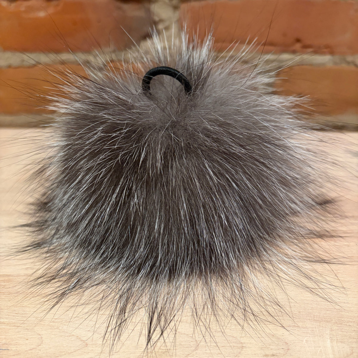 A gray silver fox fur pom-pom a loop attachment, resting on a light wood surface with a brick wall background.