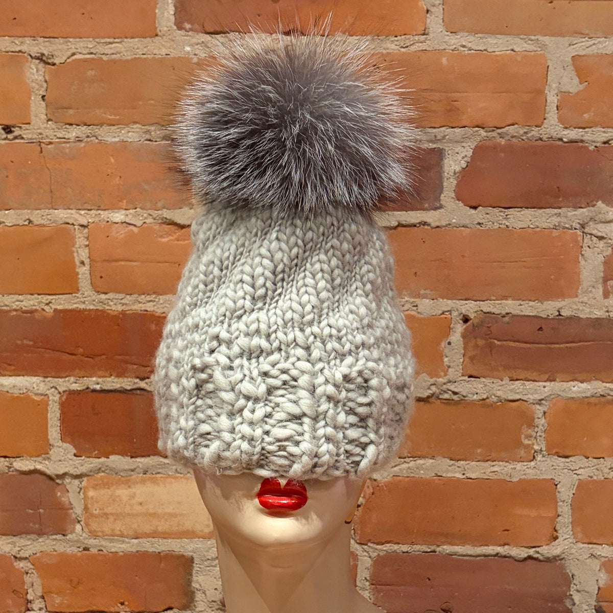A chunky knit winter beanie in a light gray color with a large fluffy gray pom-pom, displayed on a mannequin head against a red brick wall background.