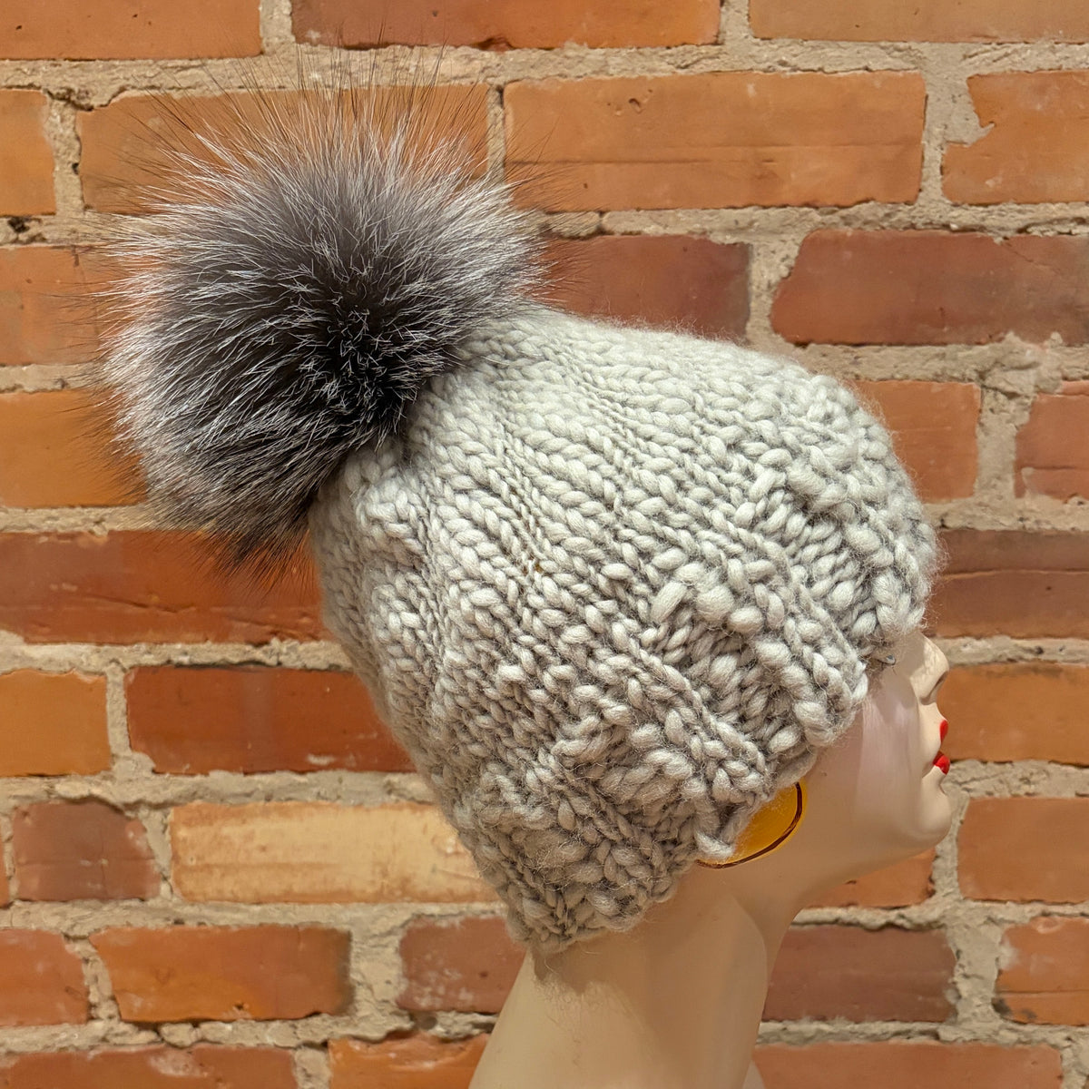 A chunky knit winter beanie in a light gray and white cable-knit pattern, featuring a large fluffy gray and white fox-fur pom-pom. The hat is displayed on a mannequin head against a red-brown brick wall background.