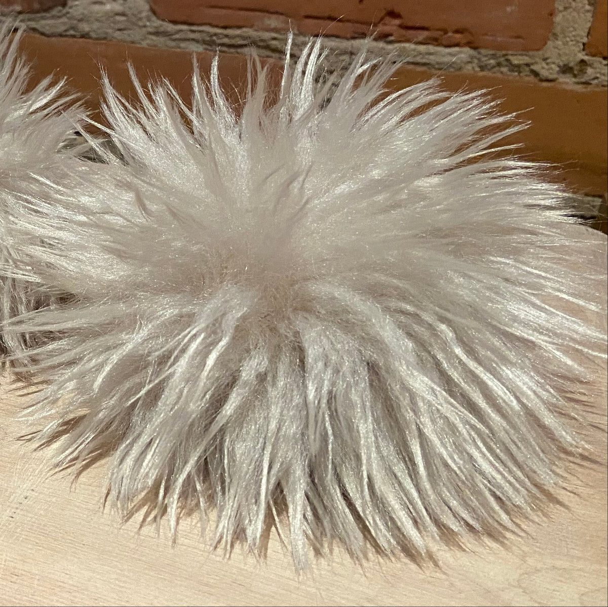 Fluffy faux-fur pom-pom in a light cream color, arranged on a light wood surface in front of a brick wall.