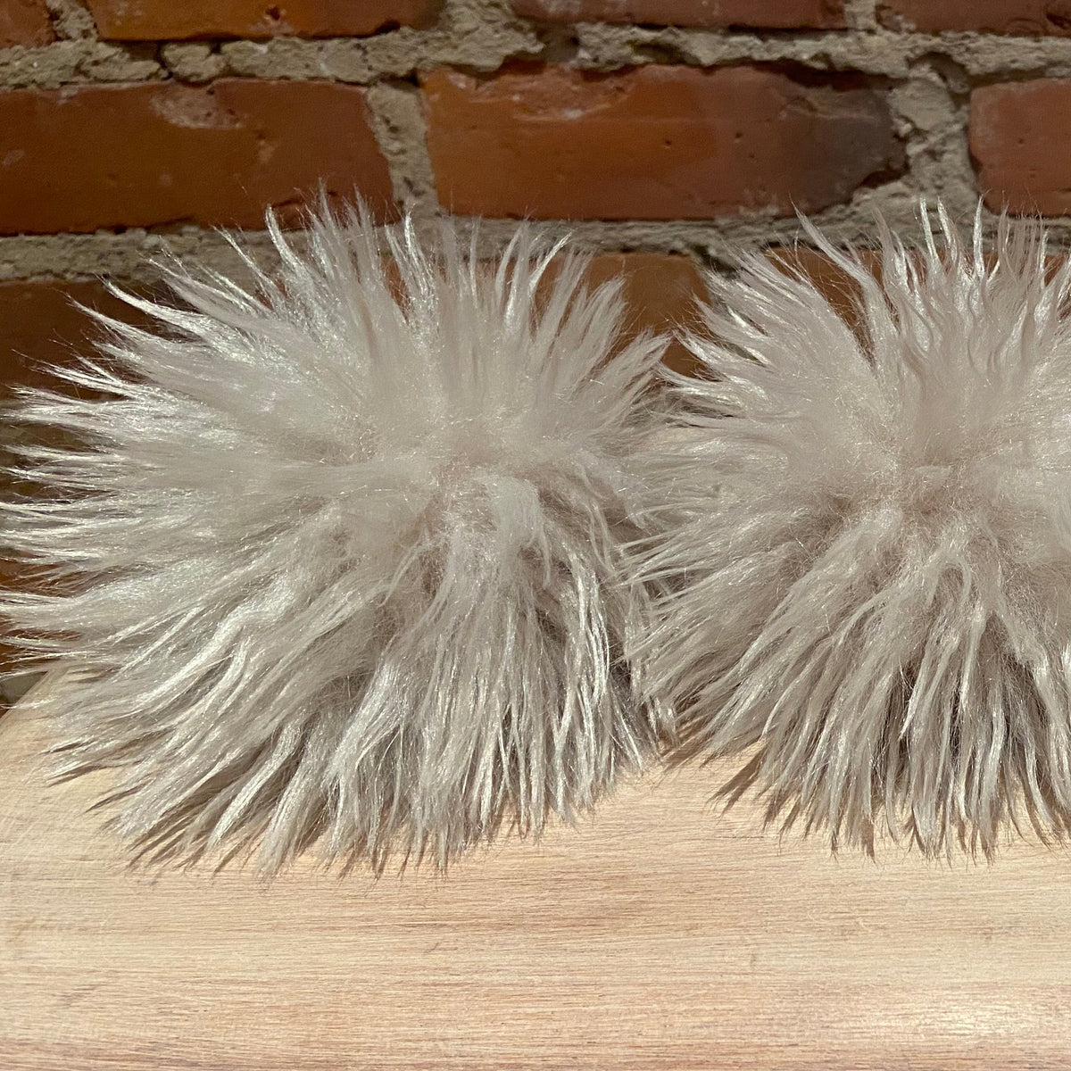 Fluffy faux-fur pom-pom decoration in a light gray color, arranged on a light wood surface with a brick wall background.