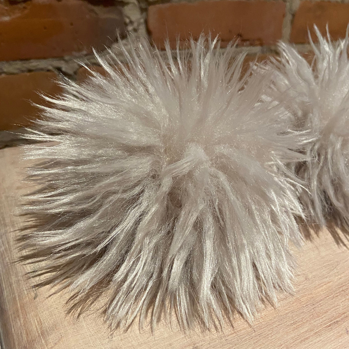 A close-up of fluffy faux fur pom-poms in a light cream or off-white color, resting on a light wood surface with a brick wall background.
