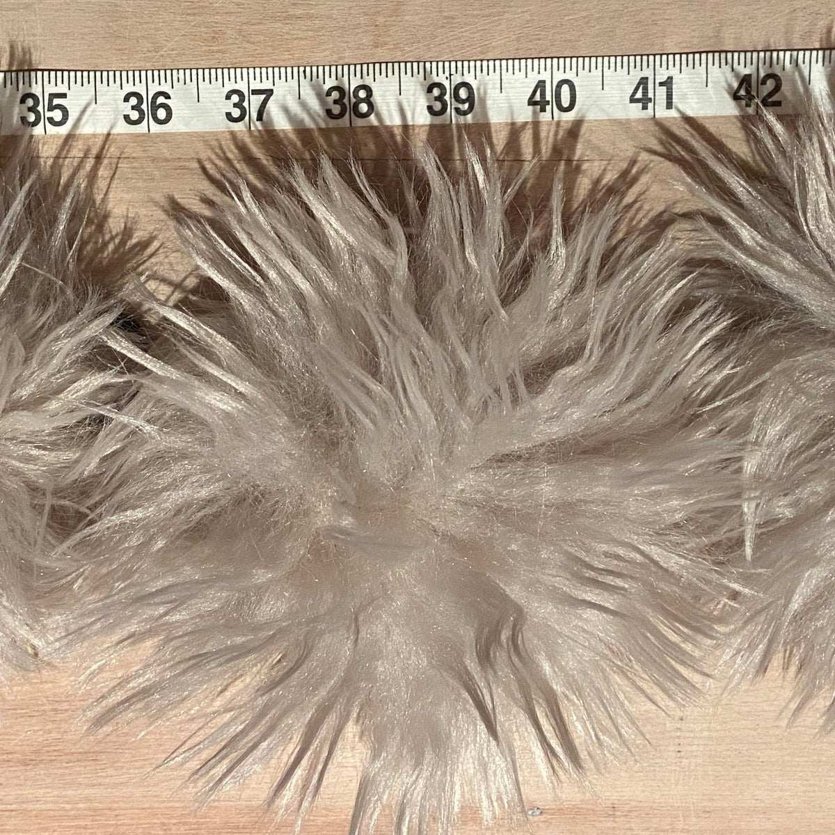 Three fluffy light gray faux poms laid out on a wooden surface with a measuring tape visible at the top for scale.