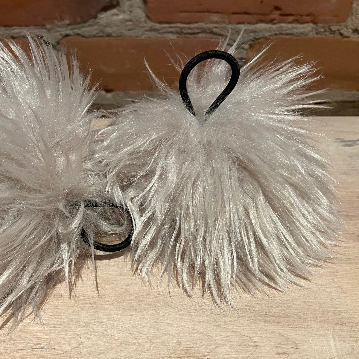 Two fluffy faux-fur pom-pom hair accessories in a light silver-gray color, each attached to a black elastic band, displayed on a light wood surface with a brick wall background.