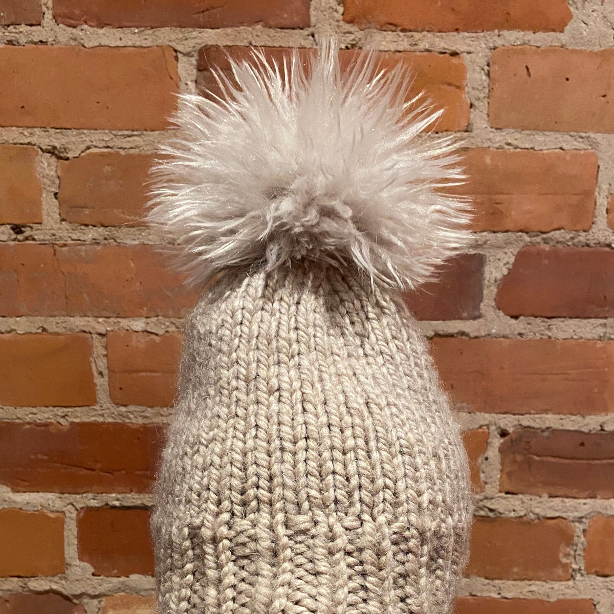 A chunky knit beanie hat in a light oatmeal beige color with a large fluffy faux-fur pom-pom on top, displayed on a mannequin head with red lips against a brick wall background.