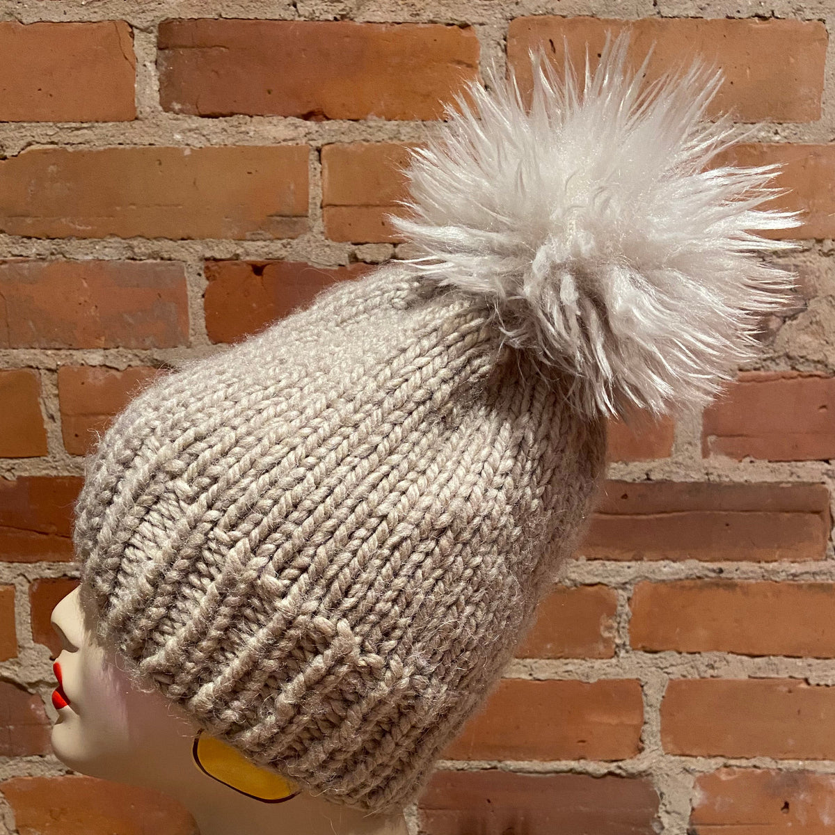 A chunky knit beanie in a light oatmeal beige color with a large fluffy white pom-pom, displayed on a mannequin head against a red brick wall background.
