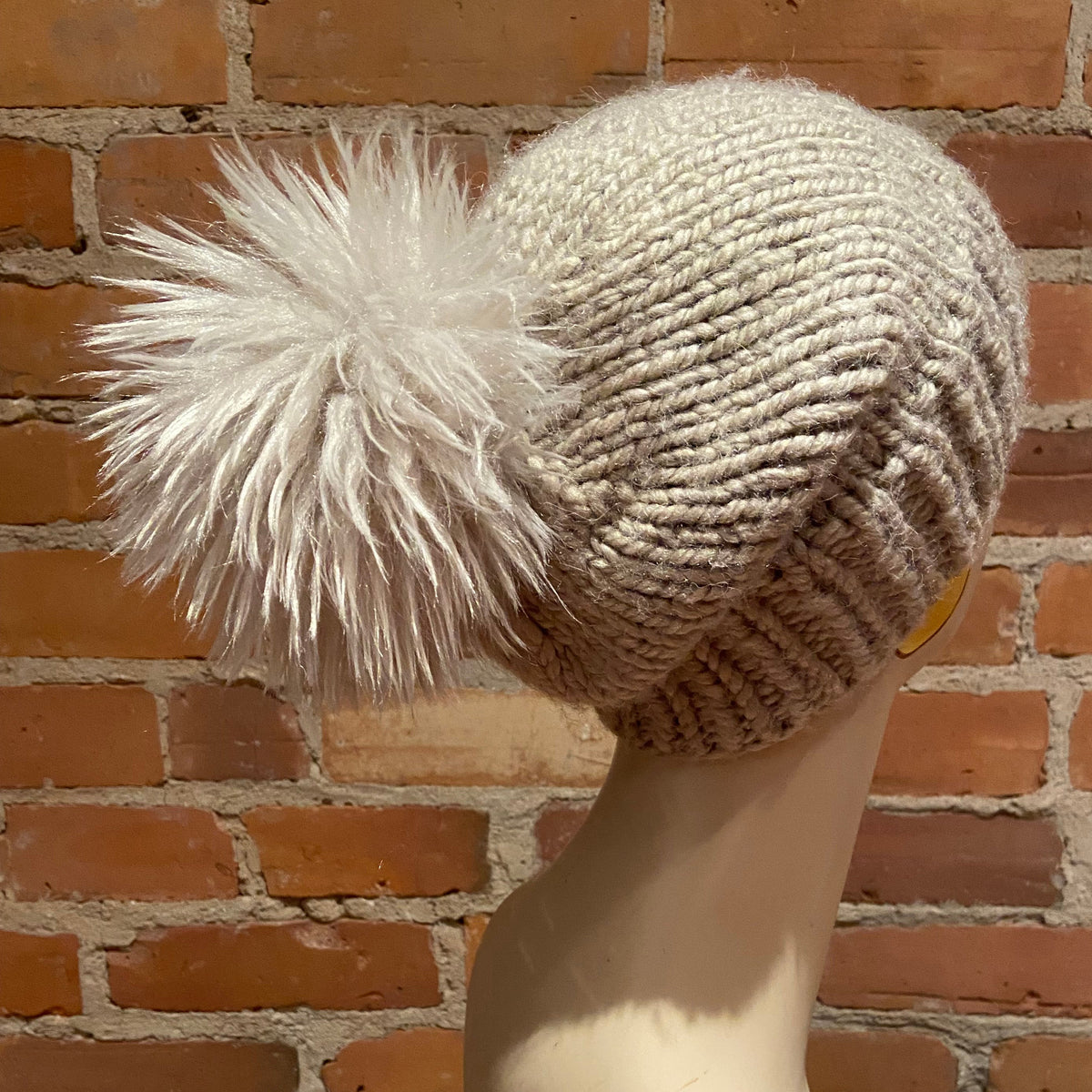 A chunky knit beanie hat in a warm oatmeal beige color with a large fluffy white faux-fur pom-pom, displayed on a mannequin head against a red brick wall background.