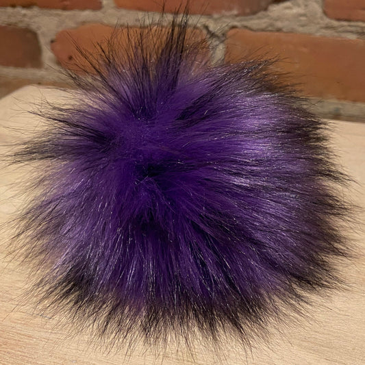 A fluffy pom-pom accessory in vibrant purple with black tips, resting on a light wood surface with a brick wall background.