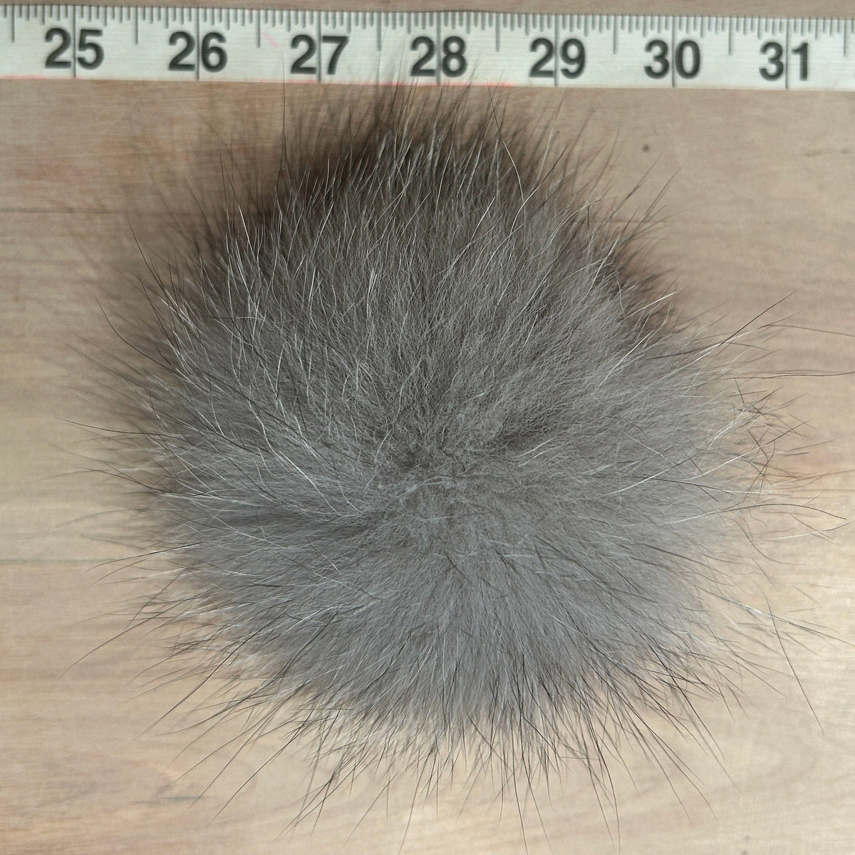 A gray fox fur pom-pom ball shown on a light wood surface with a measuring tape ruler at the top to indicate its size.