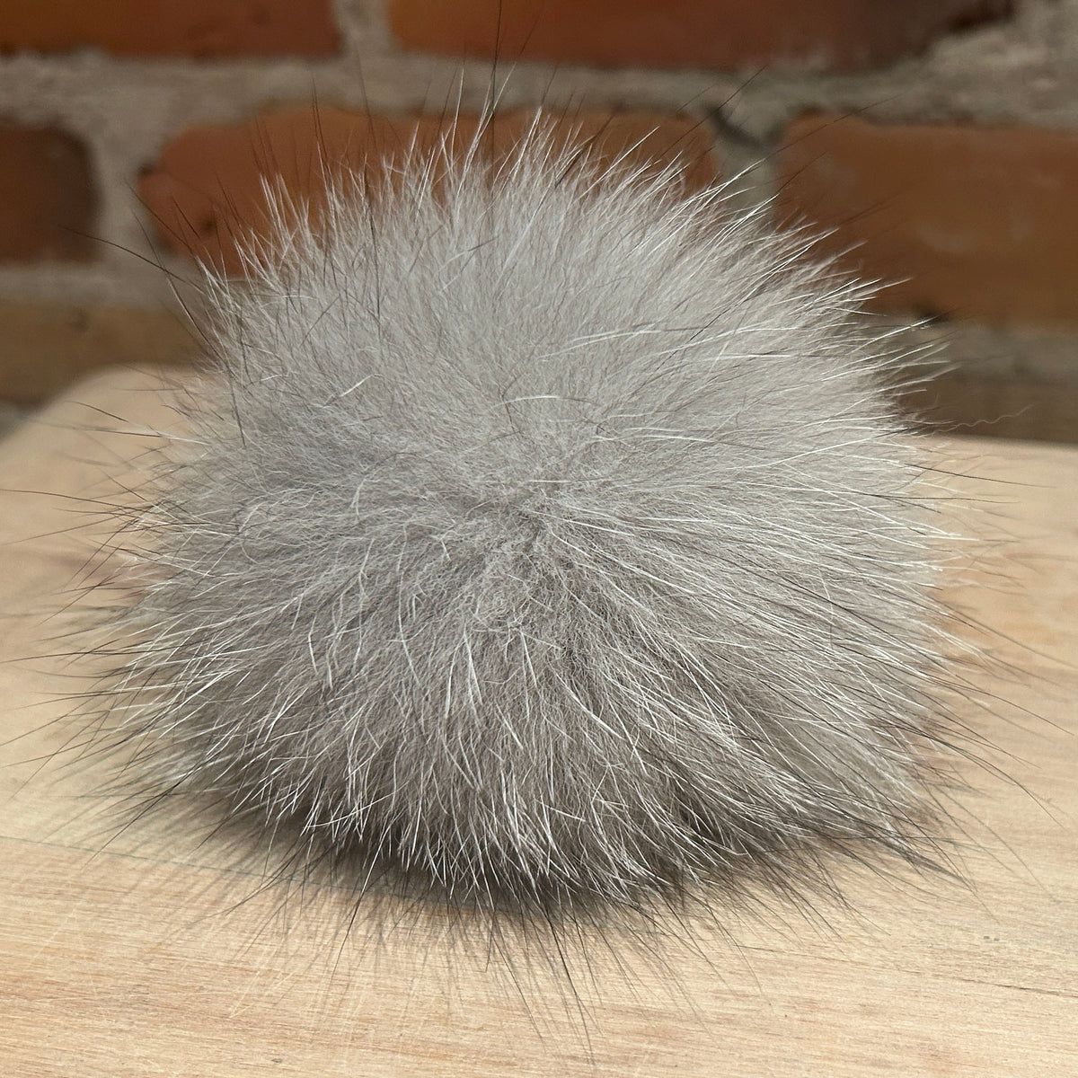 A fluffy pom-pom ball made of light gray fox fur or hair, resting on a light wood surface with a blurred brick wall background.