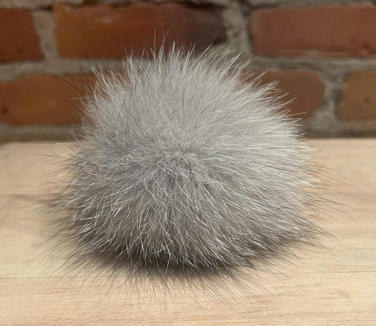 A fluffy gray fox fur pom-pom ball resting on a light wood surface with a blurred brick wall background.