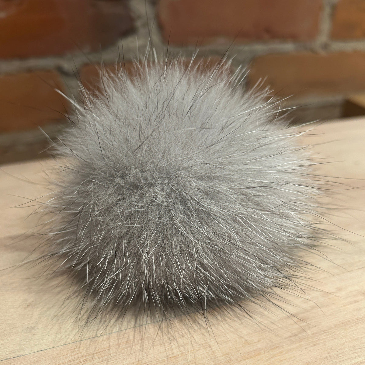 A fluffy gray fox fur pom-pom accessory resting on a light wood surface with a blurred brick wall background.