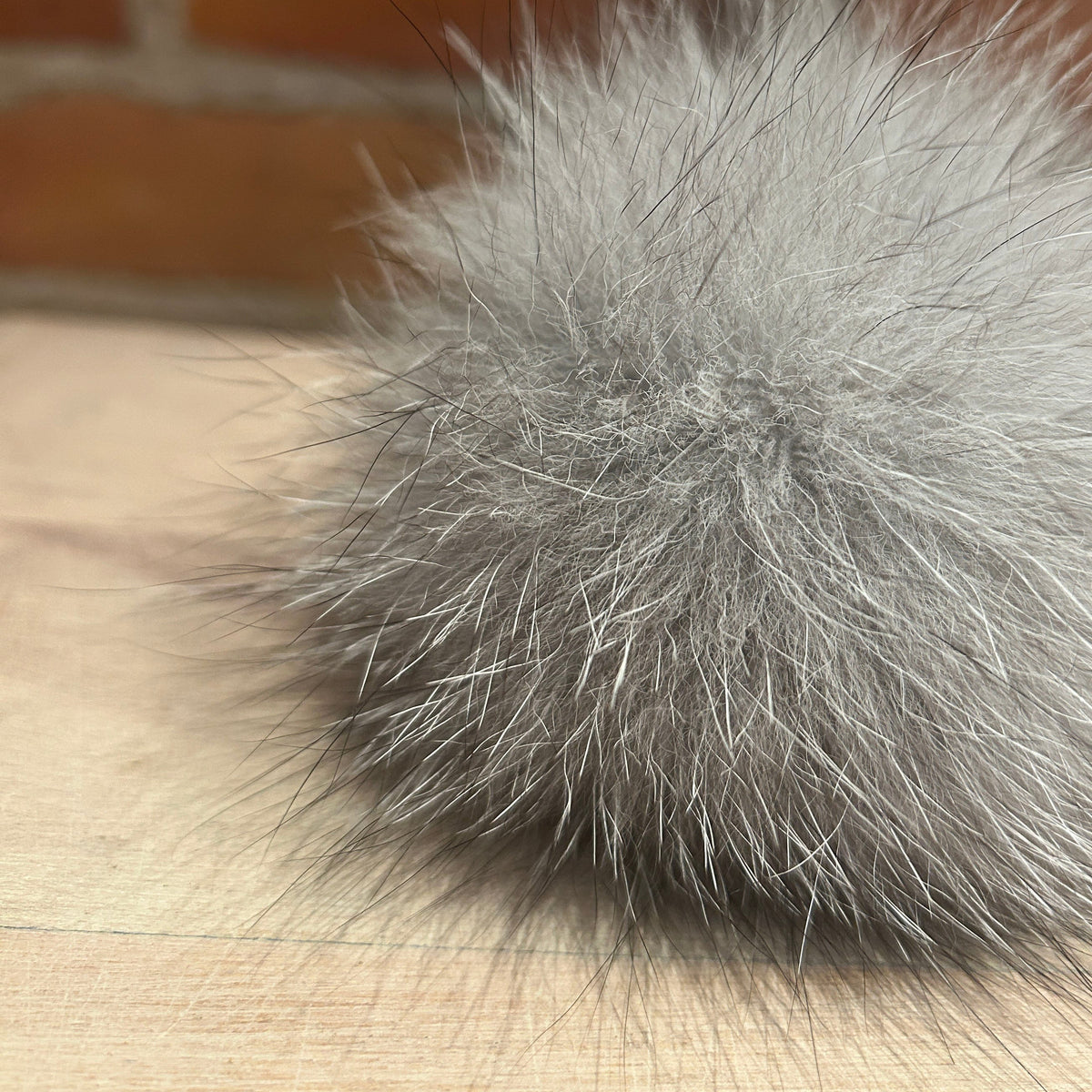 A close-up of a fluffy gray fox fur pom-pom resting on a light wood surface with a blurred brick wall background.