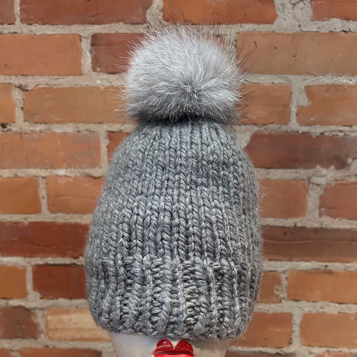 A gray ribbed knit beanie with a fluffy light gray pom-pom, displayed on a mannequin head with red lips in front of a red brick wall.