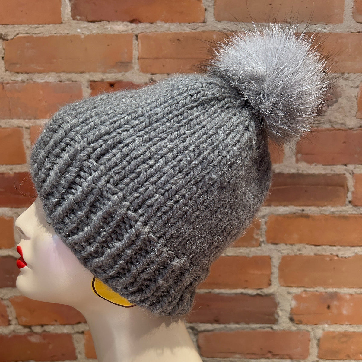 A gray ribbed knit beanie with a fluffy light gray pom-pom, displayed on a mannequin head against a red brick wall background.