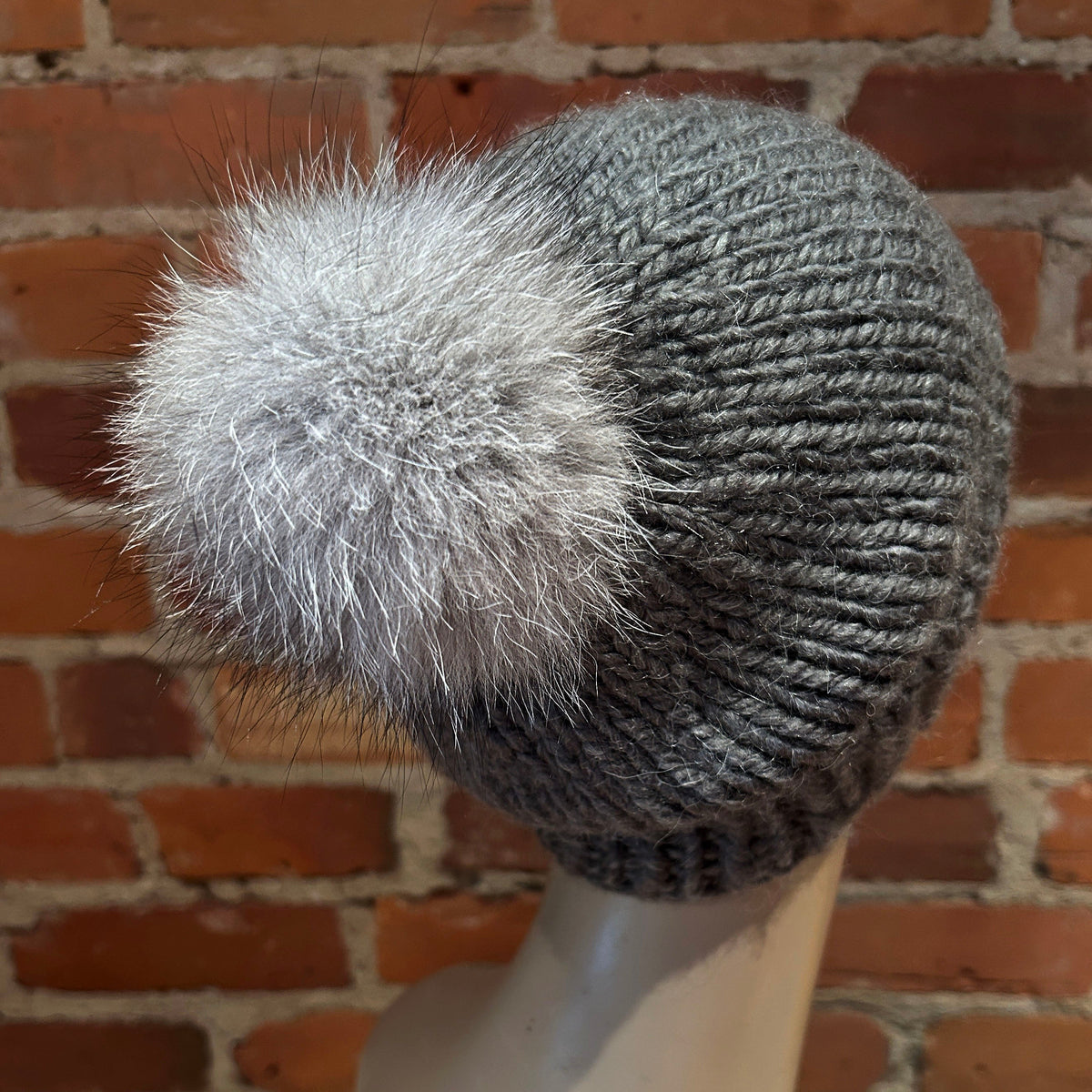A dark gray ribbed knit beanie hat with a fluffy light gray pom-pom, displayed on a mannequin head against a red brick wall background.